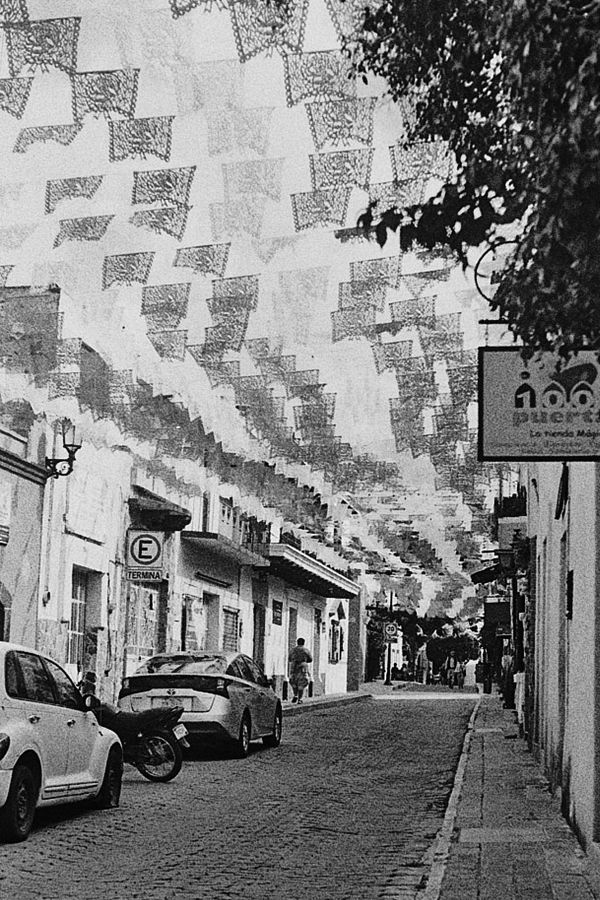 Calle angosta cubierta de papel picado, con autos estacionados