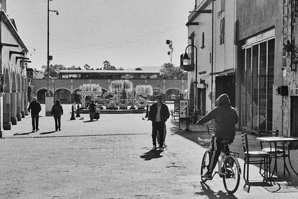 Un ciclista y peatones cruzando una calle empedrada rumbo a la plaza